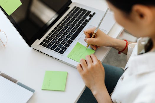 Person writing a to-do list on sticky note next to laptop for organization.